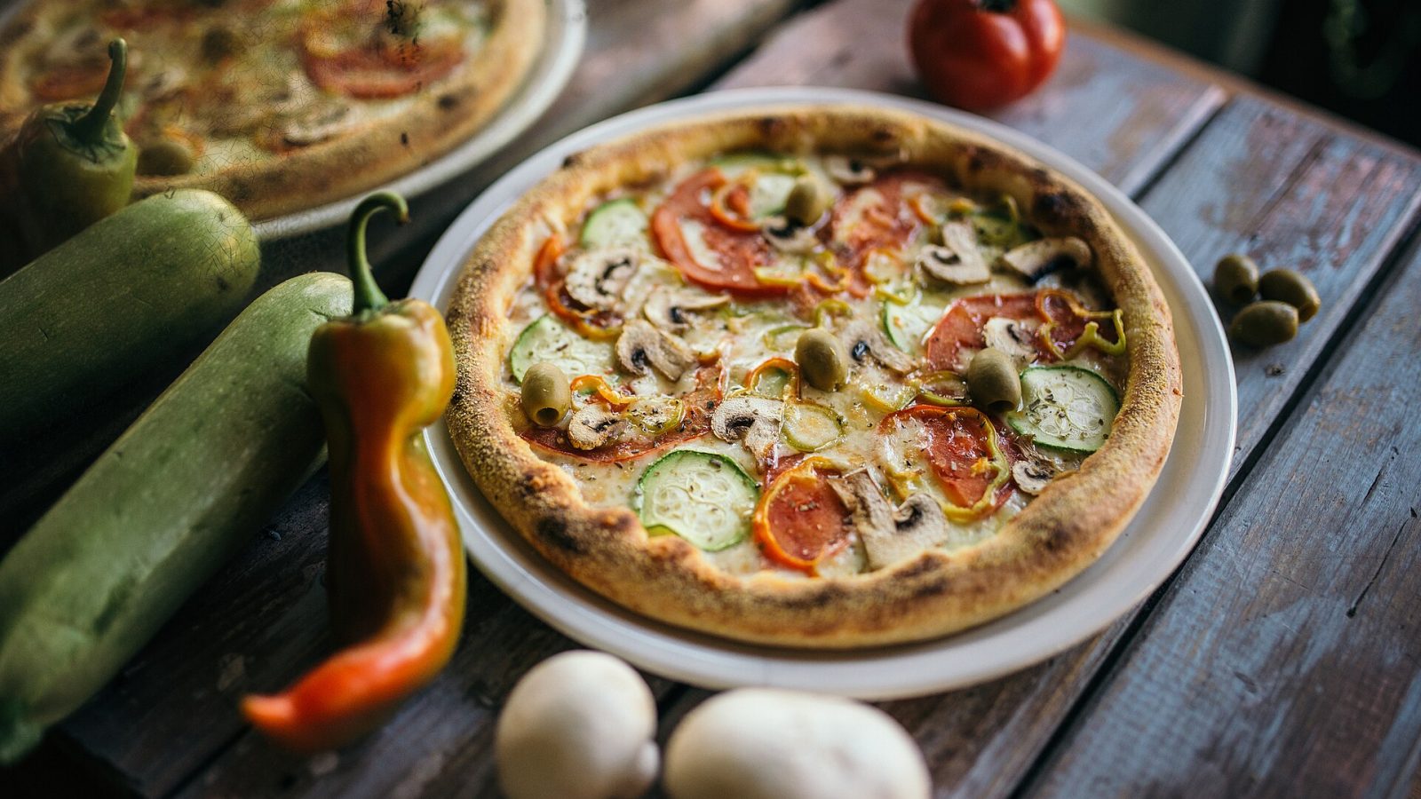 Ahşap masada kabaklı, mantarlı, domatesli ve biberli vejetaryen pizza, yanında çiğ sebzeler